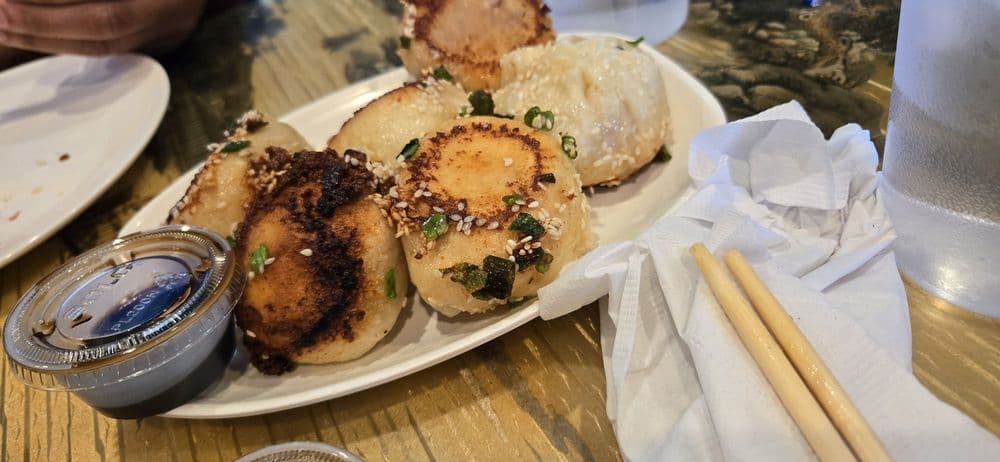 A plate of crisp-bottomed Sheng Jian Bao with sesame and scallions, beside chopsticks and dipping sauce